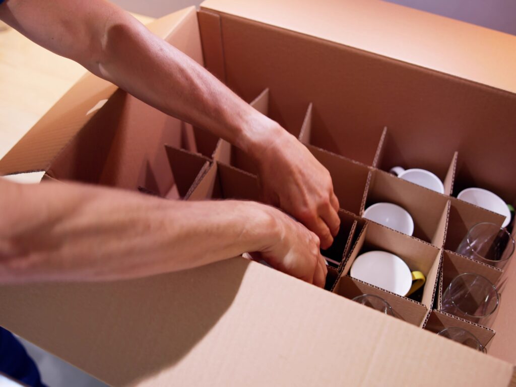 A man and van crew is packing drinking glasses and mugs into a box for moving